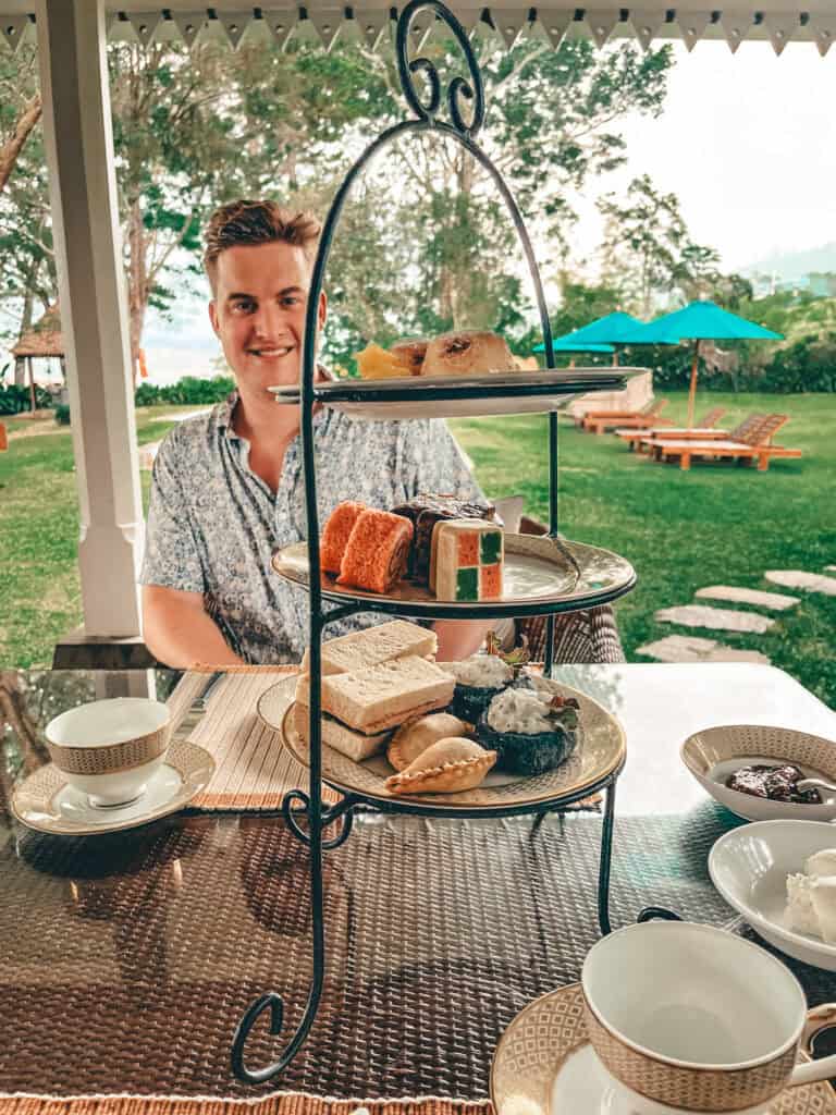 A smiling man sitting at a table set for afternoon tea, with a tiered tray displaying an assortment of cakes, sandwiches, and pastries. The table is adorned with elegant china teacups and saucers, set outdoors in a lush garden with green lawn and trees.