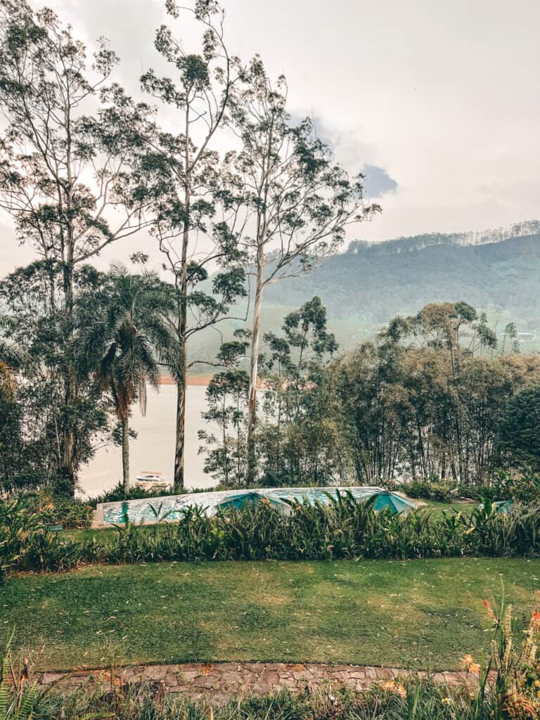 A serene garden with a lush lawn, overlooking a rectangular swimming pool surrounded by tall trees and dense foliage. The view extends to a tranquil lake and misty hills in the background, creating a peaceful and picturesque scene.