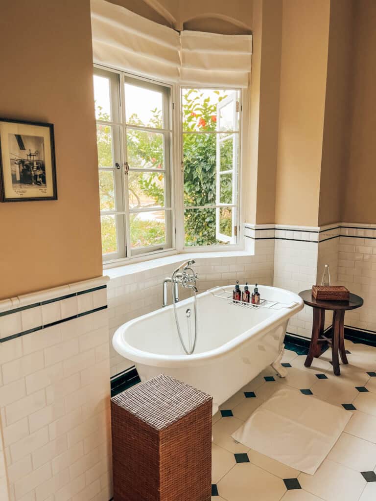 A vintage-style bathroom with a freestanding bathtub under a large window, allowing natural light to flood in. The room features beige walls, white subway tiles, and a wicker laundry basket, creating a serene and inviting atmosphere.