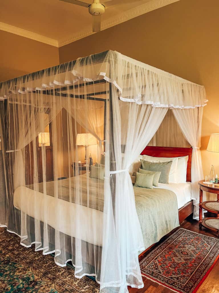 A cozy bedroom featuring a four-poster bed draped with white sheer curtains, giving a romantic and elegant touch. The bed is adorned with multiple pillows and a soft throw, while bedside tables with lamps and a patterned rug add warmth to the space.