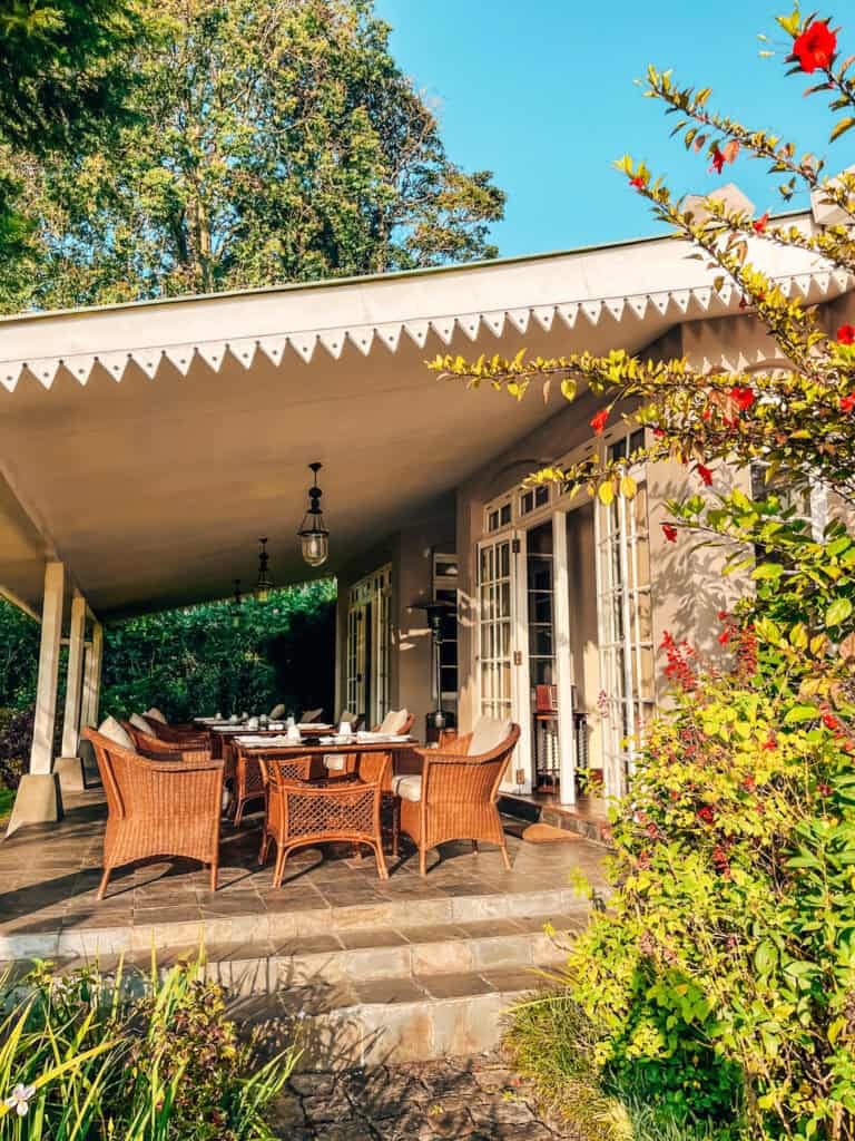 An inviting outdoor dining patio with wicker chairs and a dining table set for a meal, surrounded by lush greenery and vibrant flowers. The patio is shaded by a white awning and overlooks a serene garden, providing a cozy and picturesque dining experience.