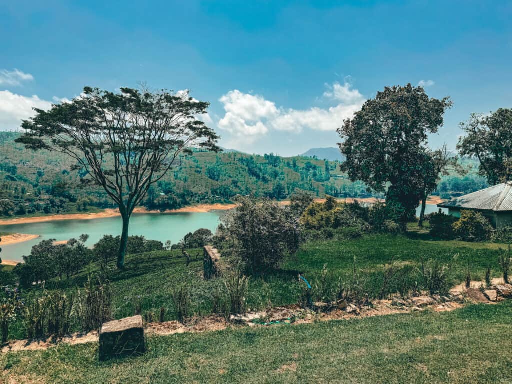 A scenic view of a lake surrounded by green hills and trees, under a bright blue sky with scattered clouds. The peaceful landscape includes a grassy area in the foreground and vibrant foliage.