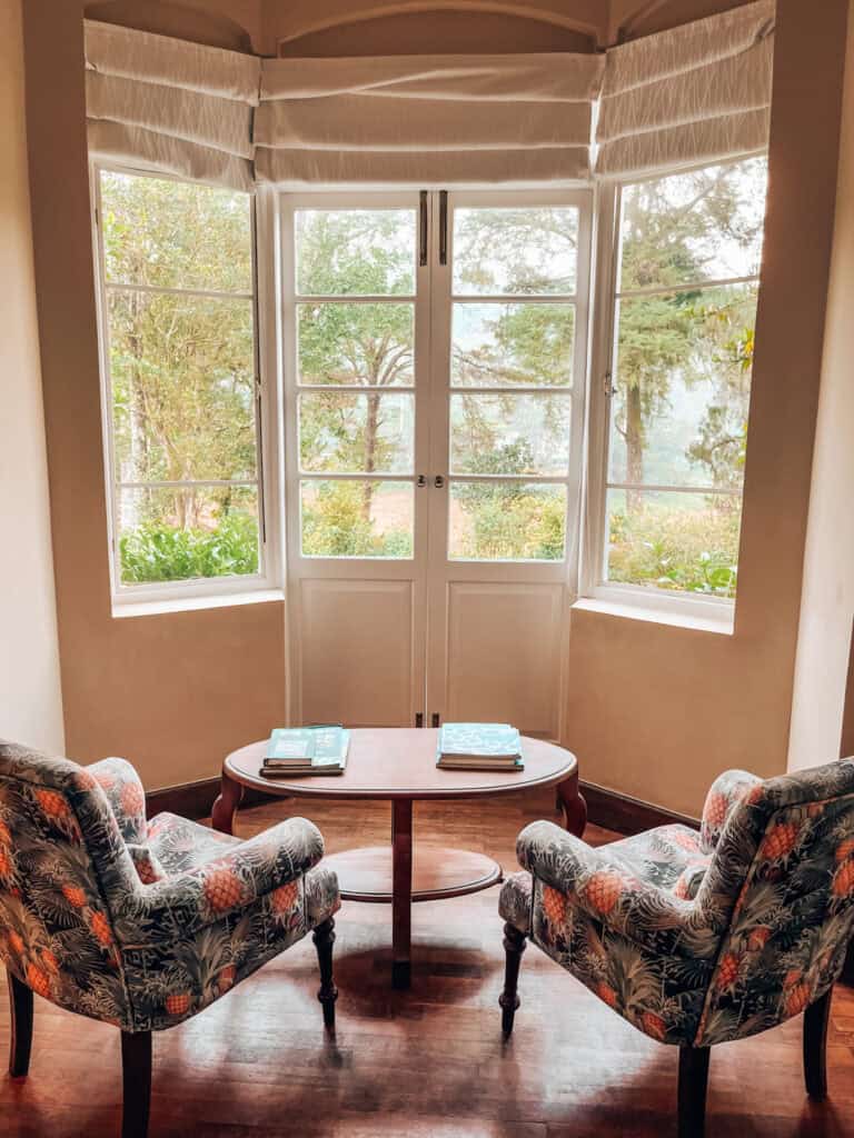 A charming sitting area with two upholstered chairs in a pineapple pattern, facing a wooden coffee table set near large, bright windows. The windows overlook a lush garden, providing a peaceful and scenic view.