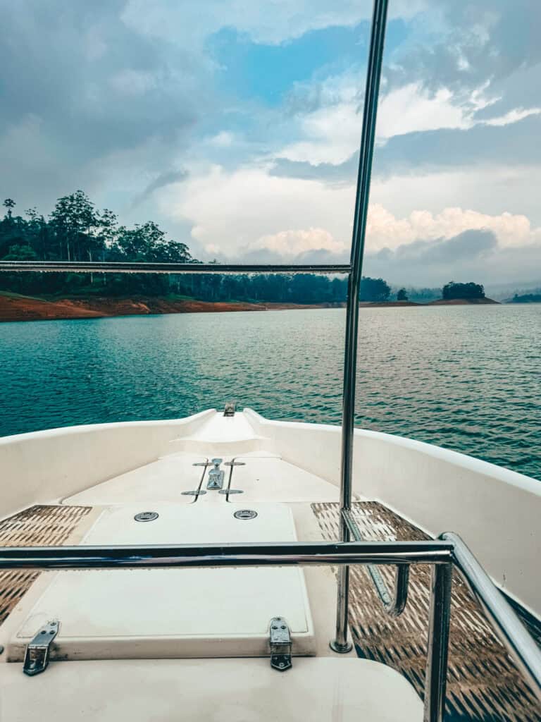 The bow of a white boat on a calm lake, with green forested hills and a cloudy sky in the distance. The image captures the sense of adventure and tranquility of a boat ride in a natural setting.