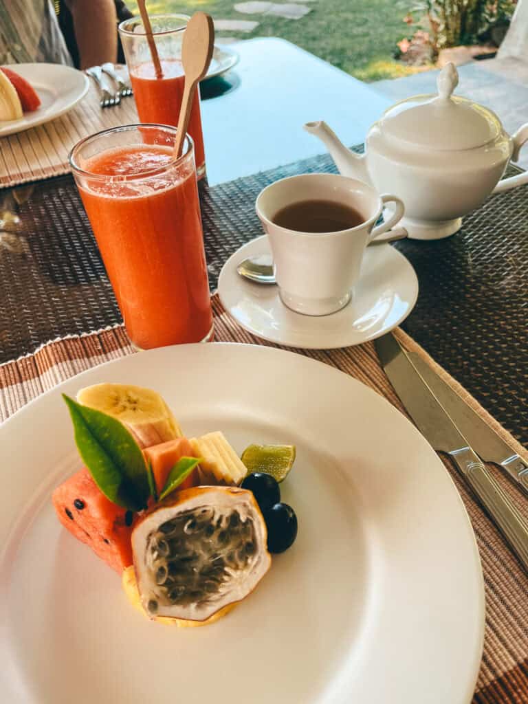 A breakfast setting featuring a white plate with a variety of fresh tropical fruits, including passion fruit, watermelon, banana, papaya, and grapes. The table also has a glass of orange juice, a cup of tea, and a teapot, arranged on a woven placemat with a garden view in the background.