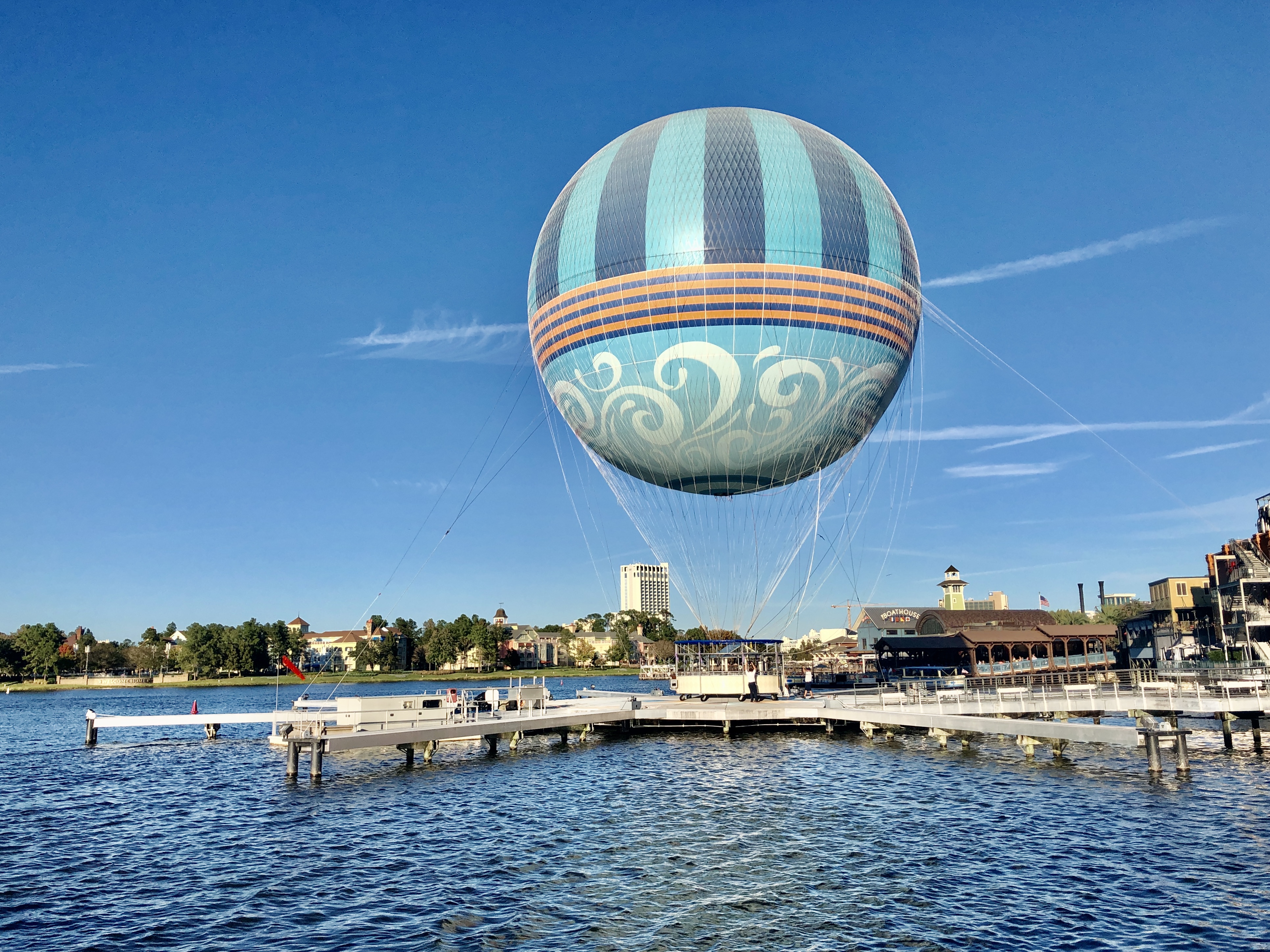 A large, tethered hot air balloon with blue and turquoise stripes and decorative swirls hovers above the water at Disney Springs in Orlando, Florida. The clear blue sky and surrounding buildings add to the picturesque scene, while the balloon prepares for its next group of riders.