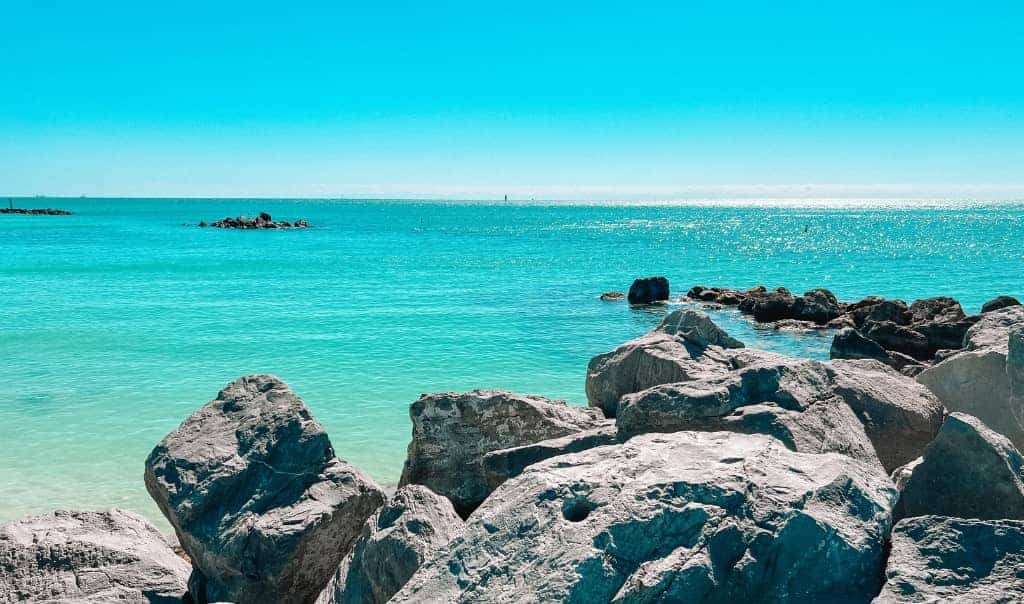 A scenic view of a serene turquoise ocean in Key West, Florida, with large, smooth rocks in the foreground and a clear blue sky overhead. The tranquil waters stretch to the horizon, reflecting the bright sunlight.