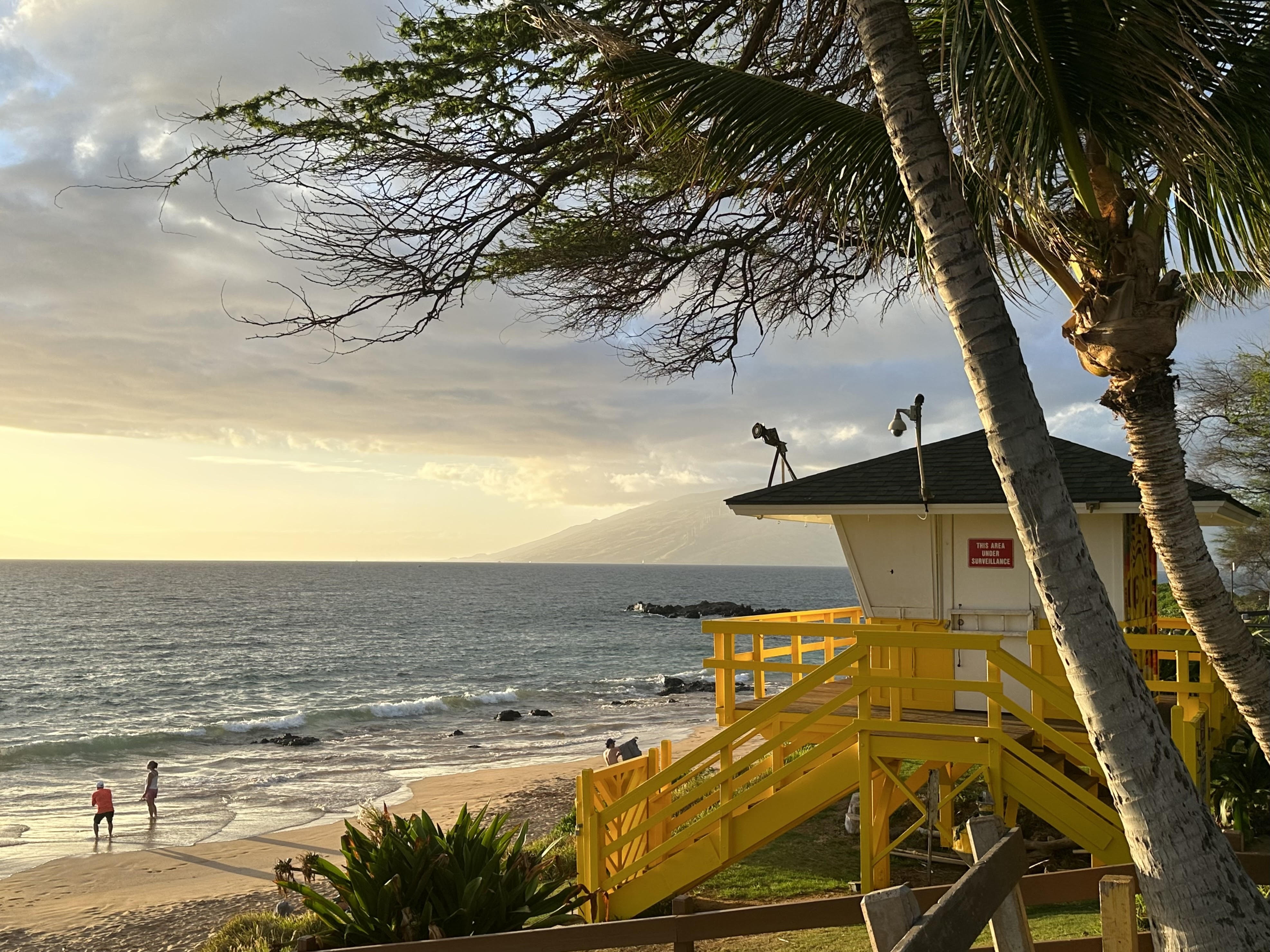 A lifeguard tower on a beach in Maui, Hawaii, framed by palm trees and overlooking the ocean at sunset. Two people stand at the water's edge, enjoying the peaceful evening scene with waves gently lapping the shore and the sky painted in soft golden hues.