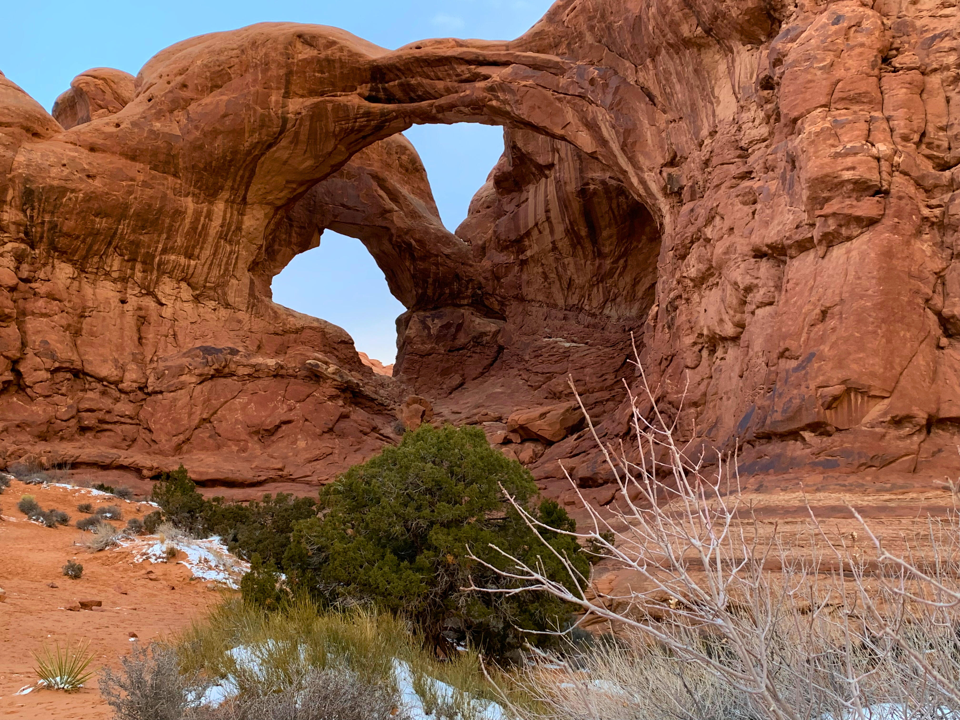 A striking natural rock formation known as the Double Arch in Arches National Park, Moab, Utah. The red sandstone arches frame the blue sky, with sparse desert vegetation and patches of snow in the foreground, highlighting the rugged beauty of the landscape.