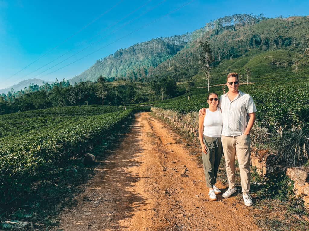 A couple standing on a dirt path surrounded by vibrant green tea plantations and rolling hills. They are smiling at the camera, enjoying the sunny day and beautiful landscape.