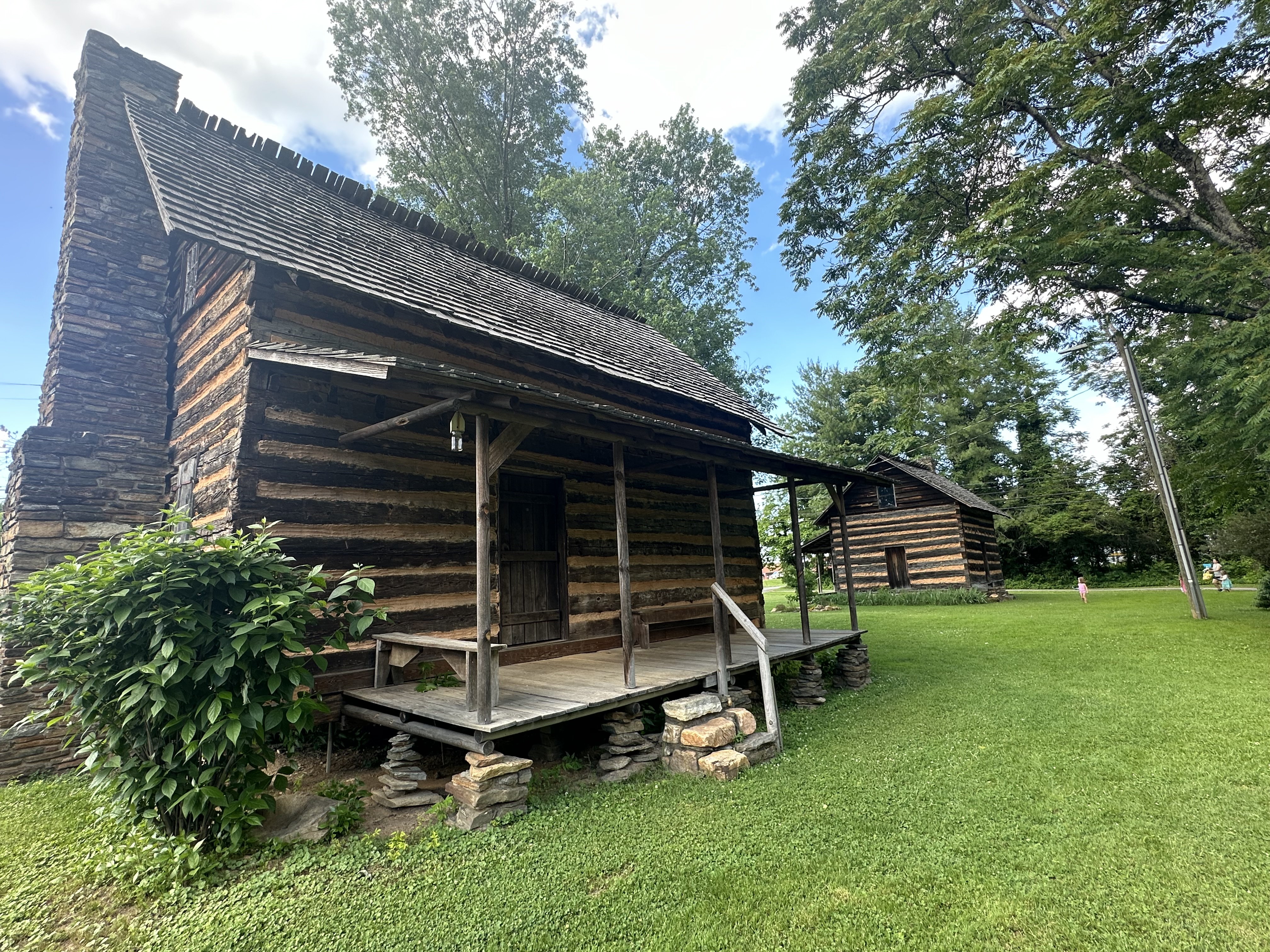 A well-preserved log cabin with a stone chimney at Old Fort, North Carolina, surrounded by lush green grass and tall trees. Another similar cabin is visible in the background, adding to the historical charm of the scene under a partly cloudy sky.