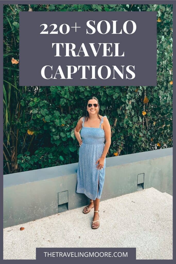 This image features a woman wearing a light blue sundress and sandals, standing in front of a green leafy background. At the top, there is large text that reads, "220+ Solo Travel Captions." At the bottom, the website "thetravelingmoore.com" is displayed. This image appears to be a promotional graphic for a blog post offering travel-related captions.