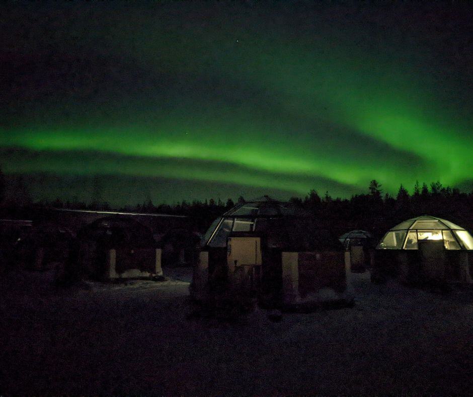 This image captures the mesmerizing Northern Lights (Aurora Borealis) glowing across the dark night sky in Rovaniemi, Finland. In the foreground, there are several glass igloo-style cabins, softly illuminated from within, allowing guests to experience the natural wonder while staying warm inside. The green hues of the Northern Lights stretch across the sky, creating a magical and serene atmosphere in this remote winter landscape.