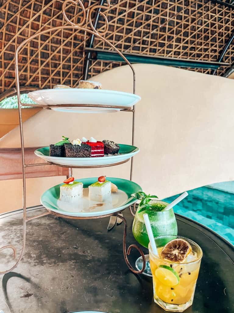 A three-tiered dessert tray with various sweets including chocolate cake, cheesecake bites, and scones, sits on a table next to two refreshing cocktails—one green with mint and one yellow with lemon and passion fruit. The background features a rustic woven roof, giving a tropical vibe.