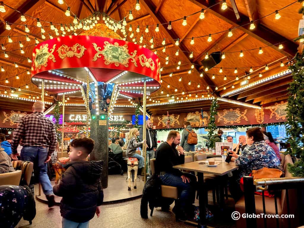 This image shows the interior of a lively café or restaurant in Amsterdam, decorated in a festive, carousel-inspired theme. The centerpiece is a large, ornate carousel canopy with string lights hanging from the wooden ceiling, creating a warm and inviting atmosphere. Patrons are seated at tables enjoying drinks and conversation, while the surroundings are adorned with holiday decorations, enhancing the cozy and cheerful ambiance of the space. A young boy is seen walking through the scene, adding a touch of liveliness to the setting.