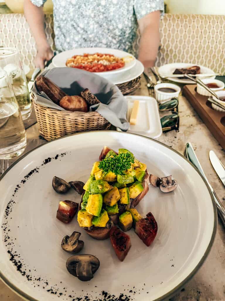 A breakfast spread featuring a plate of avocado toast topped with diced avocado, mushrooms, and roasted tomatoes, garnished with parsley. In the background, there is a basket of assorted bread, a plate of shakshuka, and small dishes of jam, butter, and other spreads, creating a vibrant and delicious meal setting.