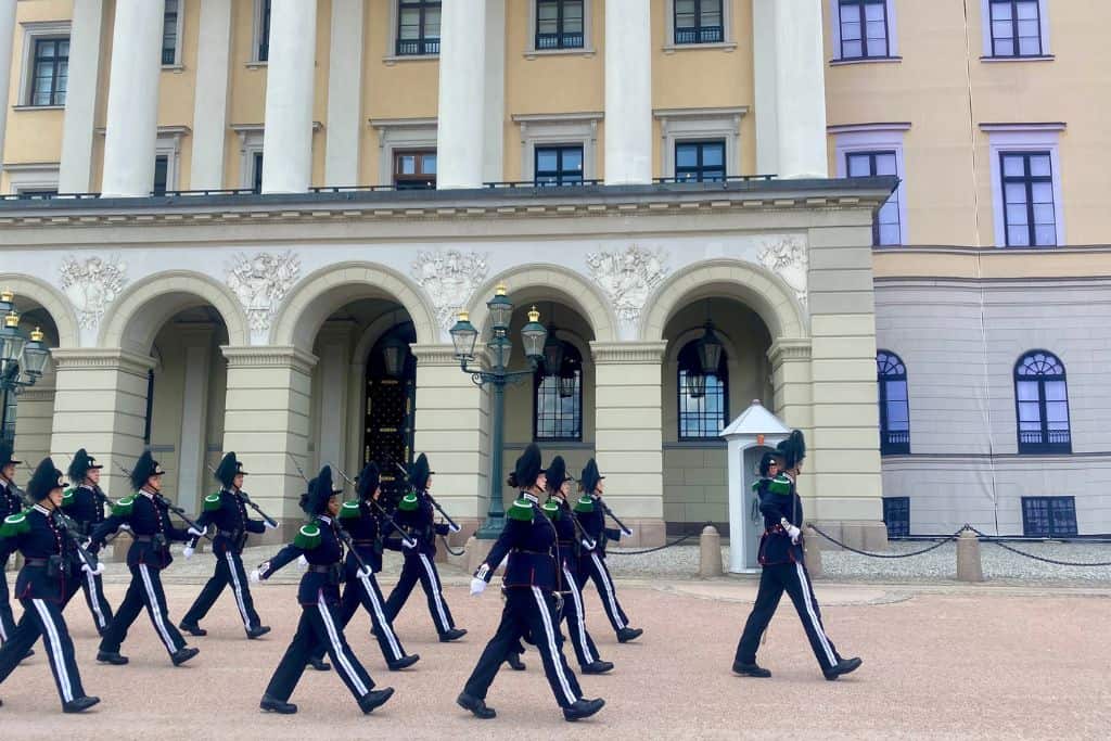 This image shows a formal changing of the guard ceremony taking place in front of a grand building, likely a royal palace in Copenhagen, Denmark. The guards, dressed in traditional dark uniforms with green accents and tall hats, are marching in sync across the courtyard in front of the palace's grand arches and columns. The guards' precision and the stately architecture in the background create an air of tradition and formality.