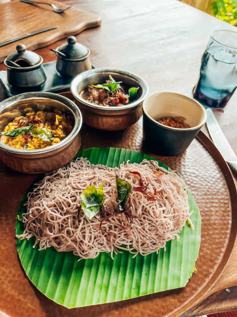 A traditional Sri Lankan breakfast featuring string hoppers (steamed rice noodles) on a banana leaf, accompanied by side dishes in small metal bowls, including dhal curry and onion sambol. The meal is served on a copper tray, with utensils and a glass of water visible in the background.