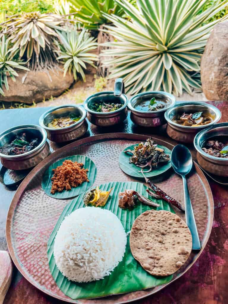 A spread of Sri Lankan curries served on a copper tray, with a mound of white rice, papadam, and various side dishes including sambol and pickles, all plated on banana leaves. The setting is tropical, with lush green plants visible in the background.