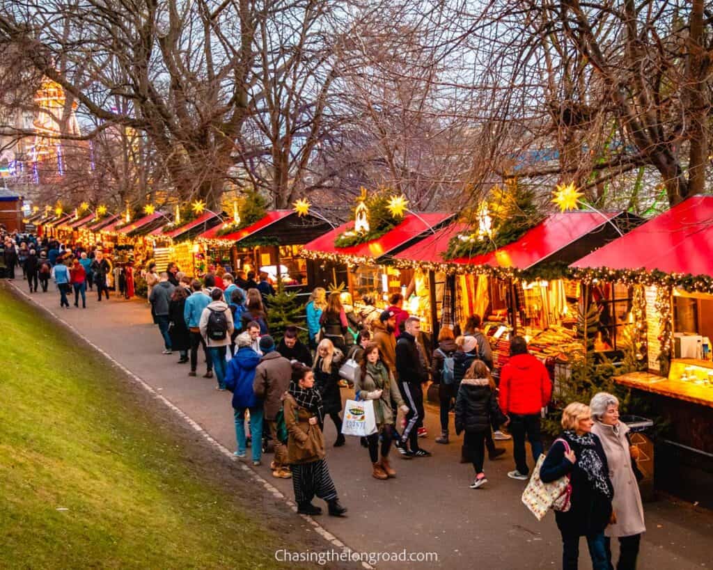 This image captures a lively Christmas market in Edinburgh, Scotland, with rows of festive stalls adorned with lights and decorations, set under bare trees. Visitors stroll along the path, browsing the various goods and enjoying the holiday atmosphere. The vibrant red roofs of the stalls and the warm glow of the lights create a cozy, inviting scene amid the cool evening.