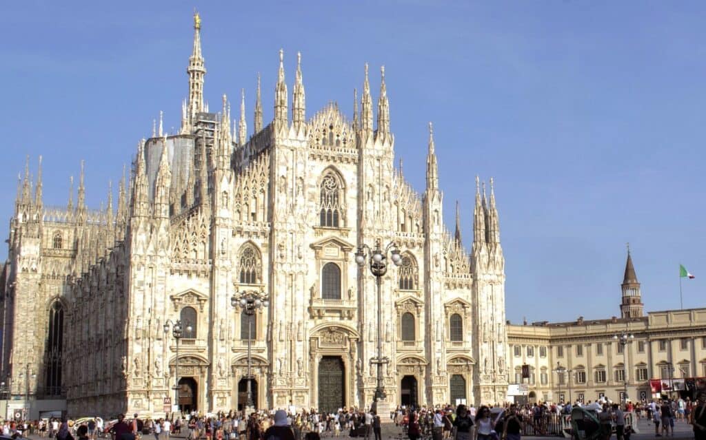 This image features the stunning Duomo di Milano (Milan Cathedral), a masterpiece of Gothic architecture, bathed in sunlight. The cathedral's intricate spires and detailed façade dominate the scene, with a crowd of people gathered in Piazza del Duomo, the large square in front of the building. The bright, clear sky complements the light-colored marble of the cathedral, while a portion of the surrounding historic buildings, including the Italian flag, is visible on the right side, adding to the grandeur of this iconic Milan landmark.