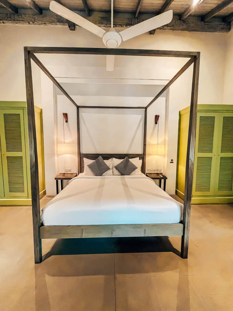 A simple, elegant four-poster bed with crisp white linens and two patterned pillows, situated between two bedside tables with modern lamps. The room features high wooden ceilings, light green closet doors, and a ceiling fan.