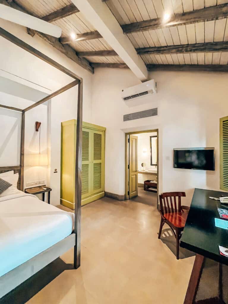  A view of the minimalist bedroom with a four-poster bed, wooden desk, and chair facing a mounted TV. The room has high ceilings with exposed beams, lime green closets, and leads into an en suite bathroom.