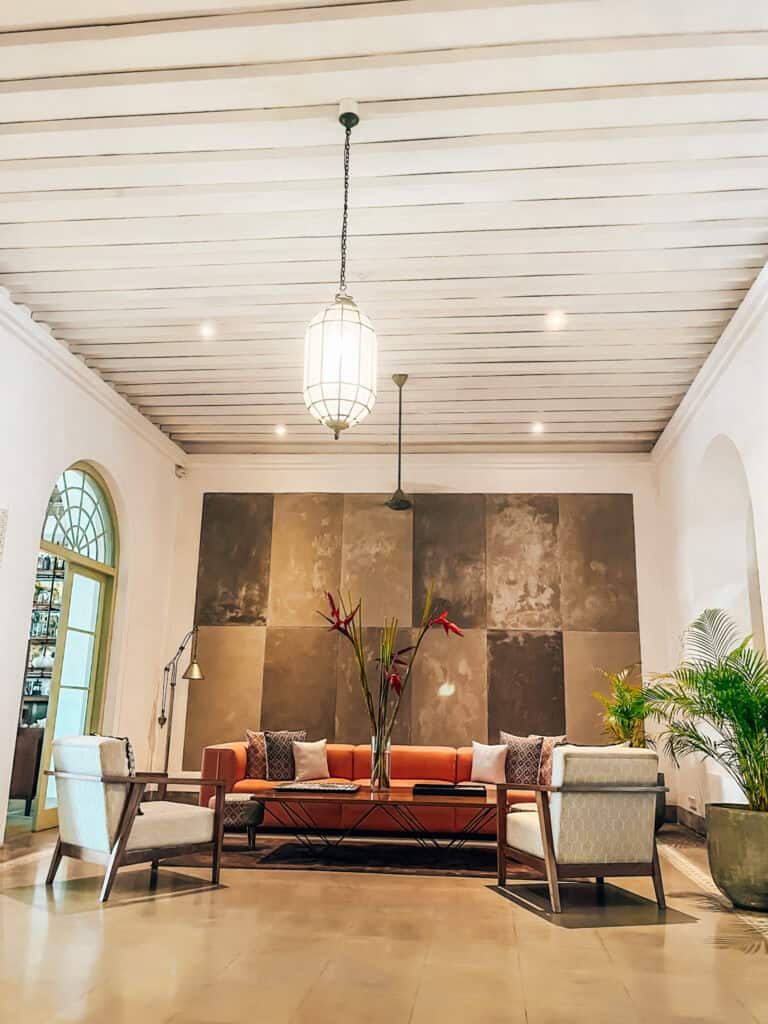 A stylish lobby featuring a bright orange sofa, flanked by two patterned armchairs, and a sleek black coffee table adorned with tall tropical flowers. The space is framed by a textured accent wall and illuminated by large, hanging lantern-style lights. Lush green plants add a natural touch to the modern decor.