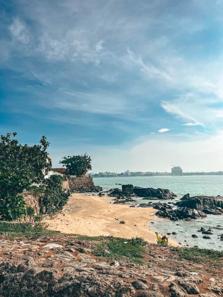 A small, serene beach with golden sand and scattered rocks, bordered by a stone wall and green foliage. The ocean is calm, and the skyline of Galle is visible in the distance under a clear blue sky.