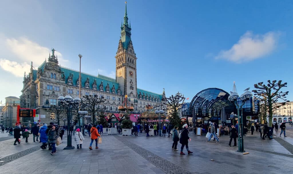 This image captures the bustling scene in front of the Hamburg City Hall (Rathaus) in Germany, with its distinctive neo-Renaissance architecture and tall clock tower dominating the view. The square in front of the Rathaus is filled with people, some visiting a small Christmas market set up with festive stalls and a carousel. The bright blue sky and the modern glass structure nearby contrast with the historic building, blending old and new elements in this urban landscape.