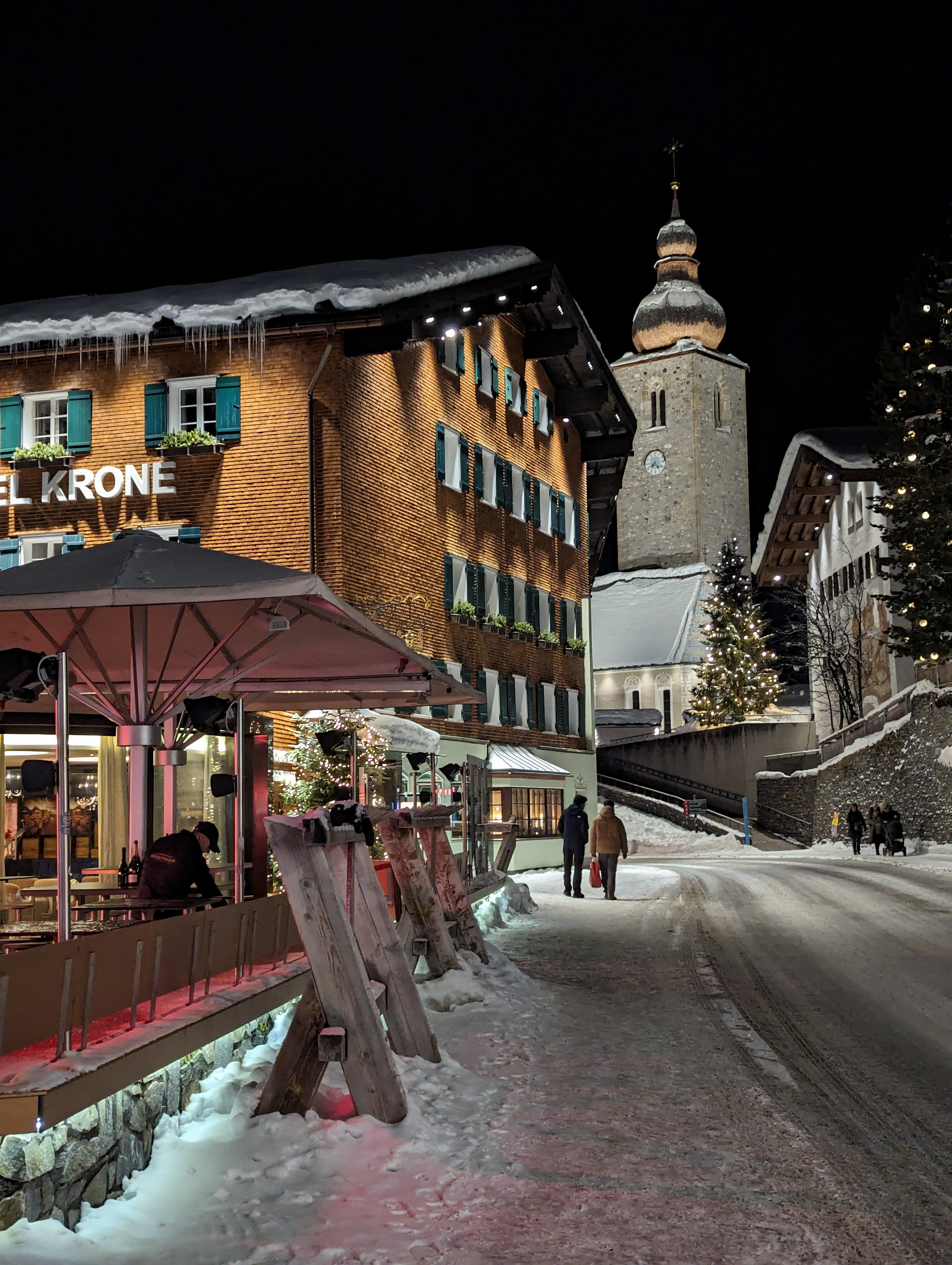 This image shows a charming winter night scene in Lech, Austria, with snow-covered streets and buildings. A cozy, well-lit hotel named "Hotel Krone" is in the foreground, its exterior adorned with icicles and soft lighting. In the background, a beautifully lit church with a traditional onion-shaped dome adds a picturesque touch to the alpine village. People are seen walking along the snowy road, enjoying the peaceful and festive atmosphere, with Christmas trees and lights adding to the seasonal charm.