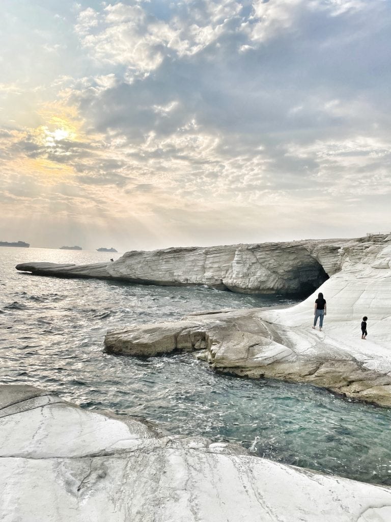 This image depicts a coastal scene in Limassol, Cyprus, featuring striking white rock formations that meet the blue waters of the Mediterranean Sea. The sky is partly cloudy with the sun peeking through, casting a soft glow over the landscape. In the foreground, two figures—a person and a child—stand on the rocks, gazing out towards the sea, adding a sense of scale and tranquility to the scene.