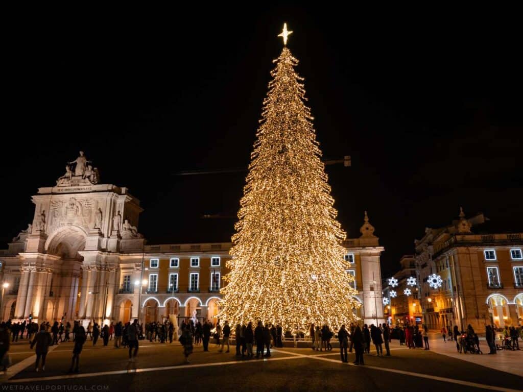 This image showcases a grand Christmas tree illuminated with thousands of lights, standing tall in Praça do Comércio, Lisbon, Portugal. The tree is the focal point of the square, with a bright star on top, drawing crowds of people who are enjoying the festive display. In the background, the iconic Arco da Rua Augusta, a large neoclassical arch, is also lit up, complementing the holiday atmosphere of the historic square. The dark night sky enhances the brilliance of the tree's lights and the surrounding decorations.