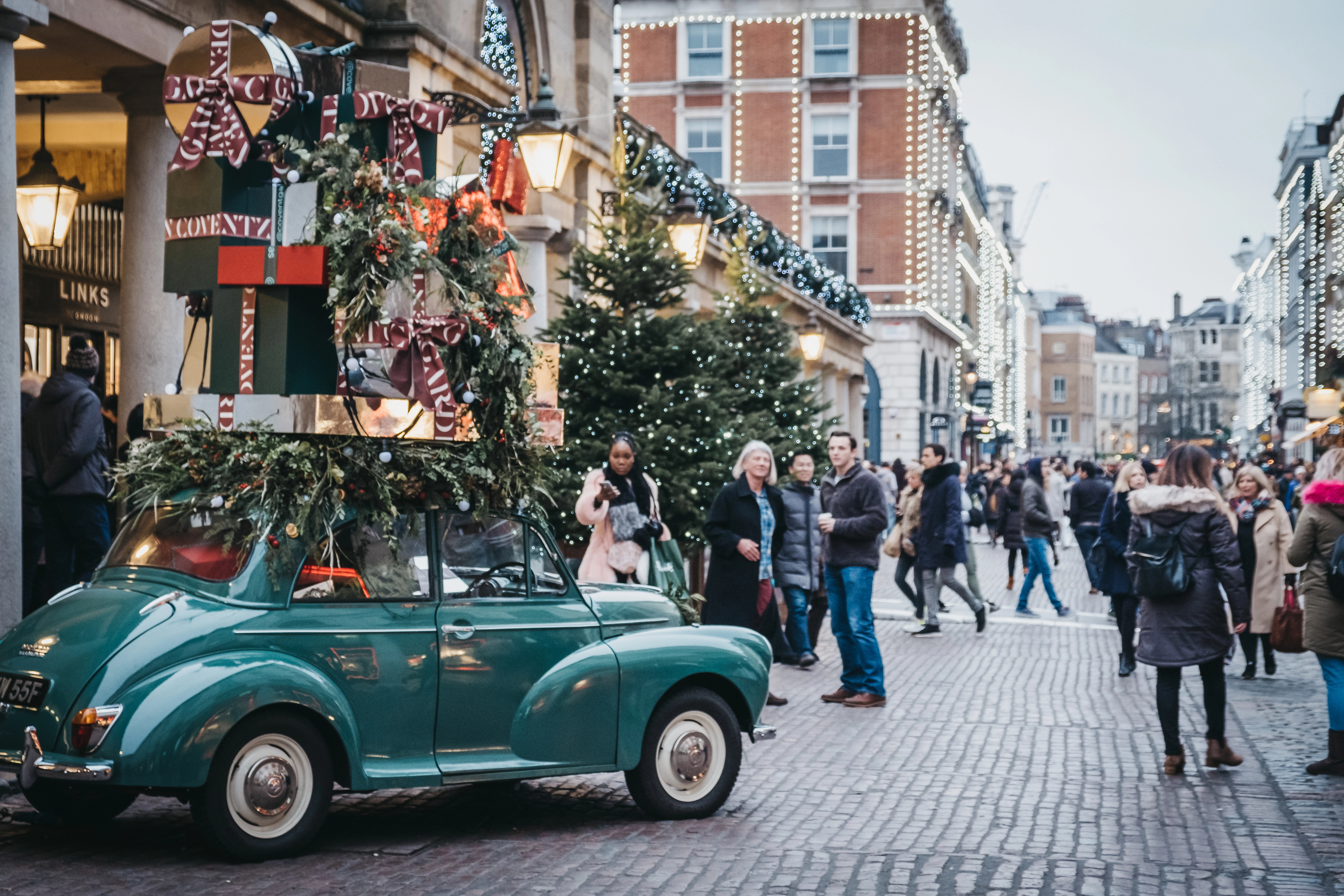 This image captures a festive scene in Covent Garden, London, during the holiday season. A vintage green car is parked on a cobblestone street, adorned with a large stack of oversized, gift-wrapped presents and greenery on its roof. The street is bustling with people, enjoying the Christmas decorations, which include beautifully lit trees and garlands strung across the buildings. The overall atmosphere is cheerful and festive, with the historic architecture of Covent Garden providing a charming backdrop to the holiday festivities.