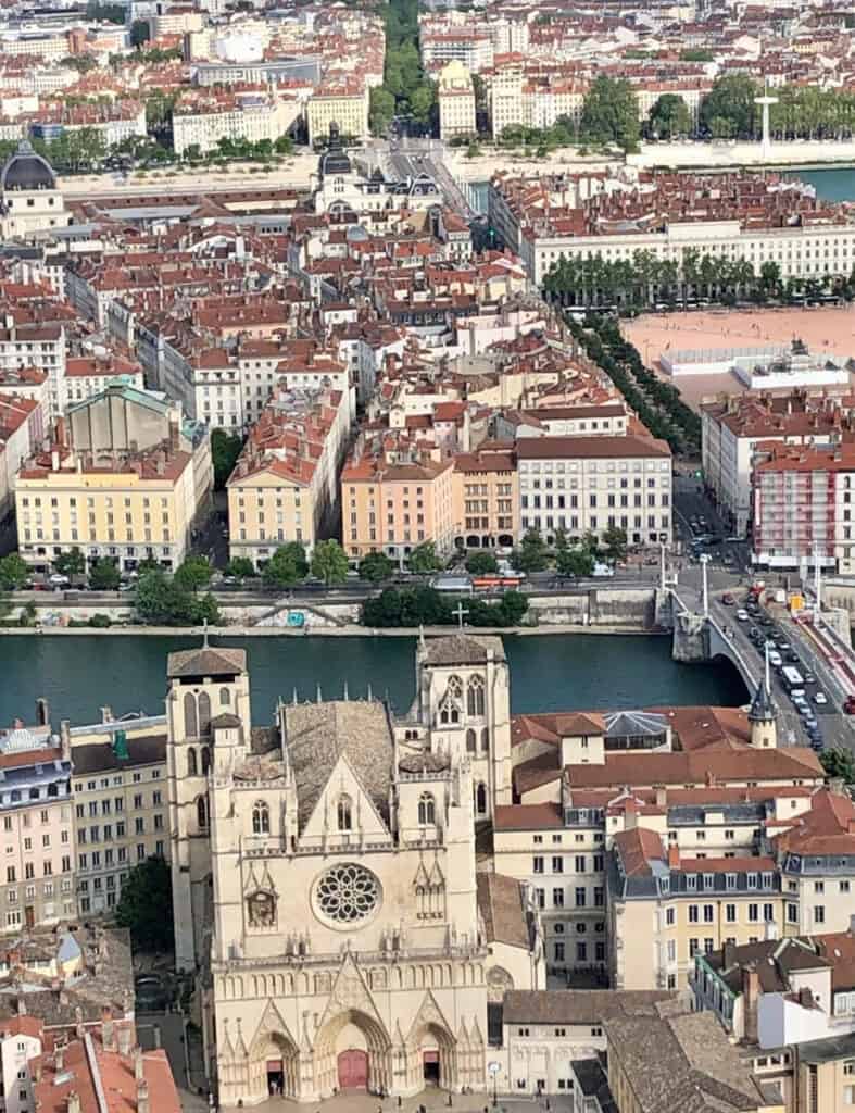 This image shows a bird's-eye view of Lyon, France, highlighting the city’s architectural landscape with rows of red-roofed buildings and the majestic Lyon Cathedral in the foreground. The Saône River runs through the middle, with a bridge connecting the historic buildings on either side. The lush greenery in the background contrasts with the dense urban environment.