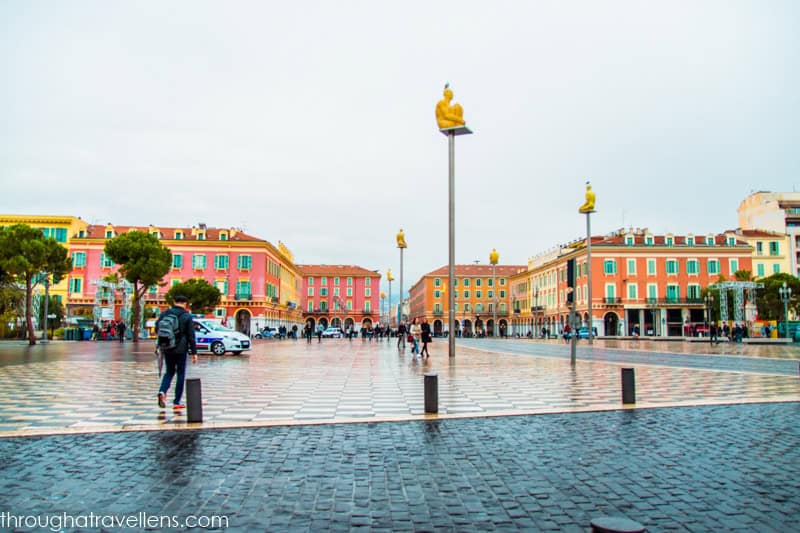 This image depicts Place Masséna, a large square in Nice, France, characterized by its distinctive black-and-white checkered pavement. The square is surrounded by colorful, historic buildings with red, pink, and yellow facades. Tall, modern sculptures of seated figures, perched atop high poles, add an artistic touch to the open space. Despite the overcast sky and wet ground, people are seen walking and enjoying the open area, giving the scene a blend of historical charm and contemporary art.