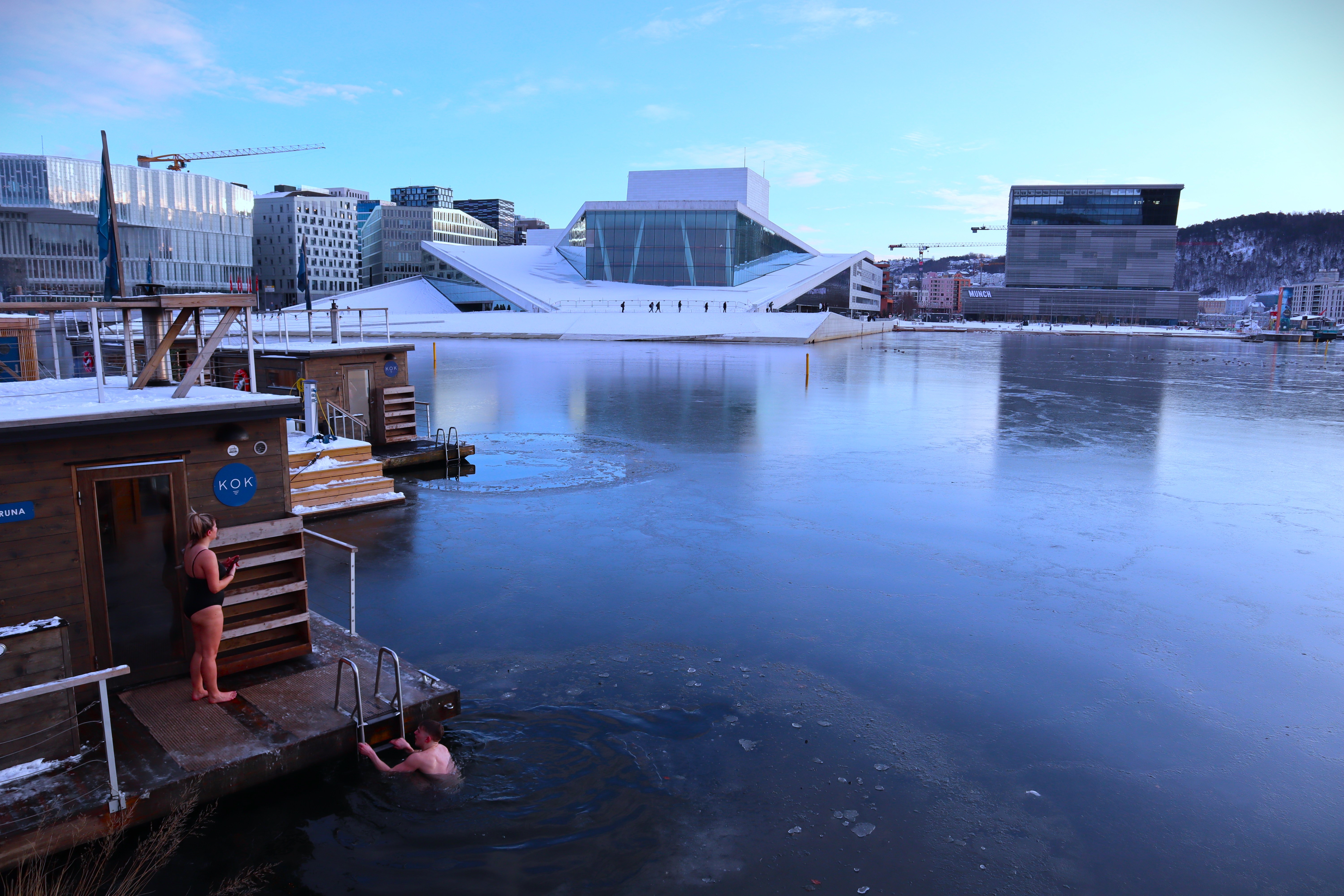 This image shows a winter scene in Oslo, Norway, featuring a person taking a dip in icy water near a floating sauna, with another person standing nearby. In the background, the modern and angular architecture of the Oslo Opera House is prominently visible across the partially frozen harbor, reflecting the cold, crisp weather. The surrounding buildings and a clear blue sky complete this striking urban winter landscape.