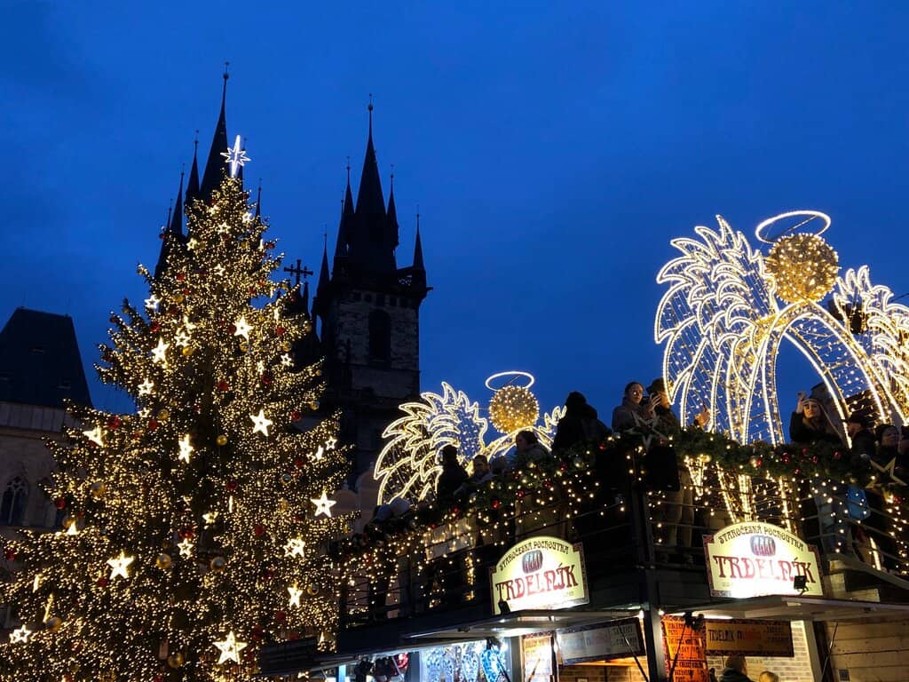 This image captures a festive Christmas market in Prague, Czech Republic, during the evening. A large Christmas tree adorned with golden lights and star ornaments stands prominently, with the spires of the Church of Our Lady before Týn rising in the background. The market stalls below, including one selling traditional 