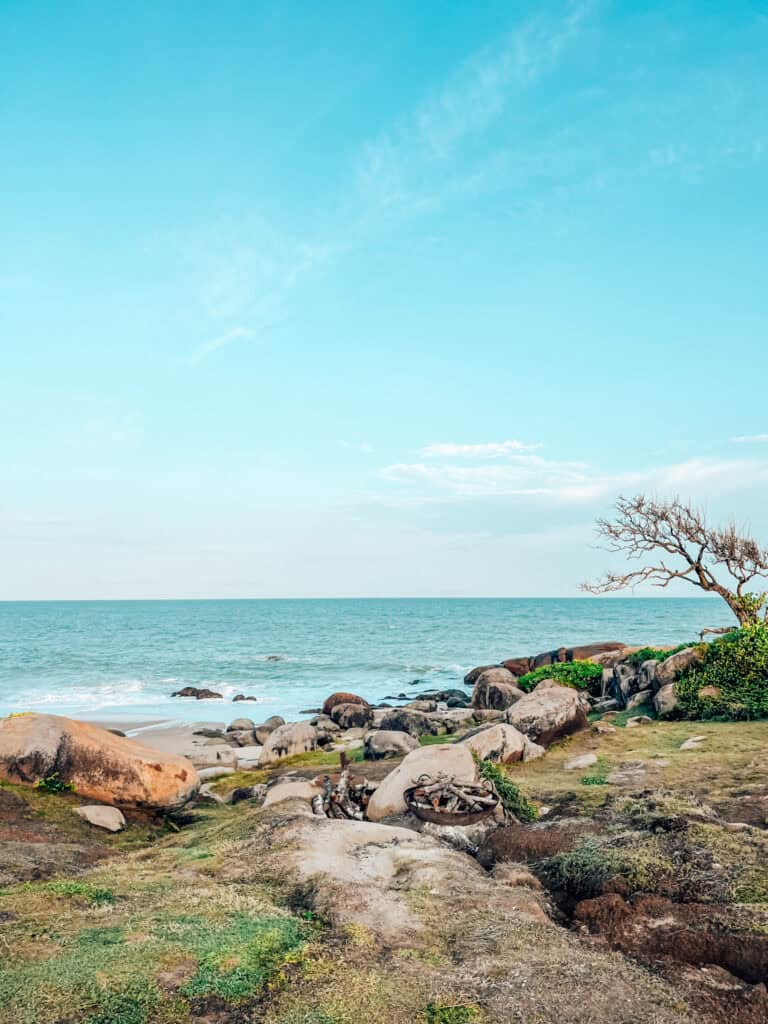 A peaceful coastline scene featuring large, smooth rocks leading down to the ocean. The sky is clear and blue, with a few scattered clouds, and a small fire pit with logs is visible in the foreground, enhancing the natural beauty of the shoreline.