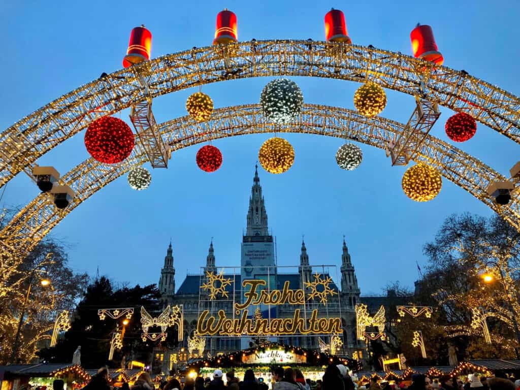 This image shows a festive Christmas market in Vienna, Austria, with a grand archway of lights and decorations framing the view. The arch is adorned with large, illuminated ornaments and candles, while the words "Frohe Weihnachten" (Merry Christmas in German) are prominently displayed in the center. In the background, the towering spires of the Vienna City Hall (Rathaus) are visible, adding a historic and majestic backdrop to the lively holiday scene below. The evening sky provides a serene contrast to the warm glow of the Christmas lights.