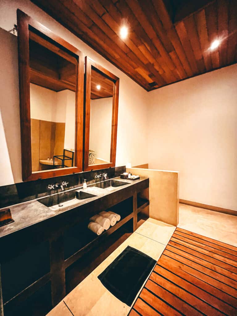 A spacious bathroom featuring a dual-sink vanity with a dark countertop, large framed mirrors, and a wooden ceiling. Towels are neatly rolled underneath the sinks, and the floor features wooden slats, adding a warm touch to the minimalist design.