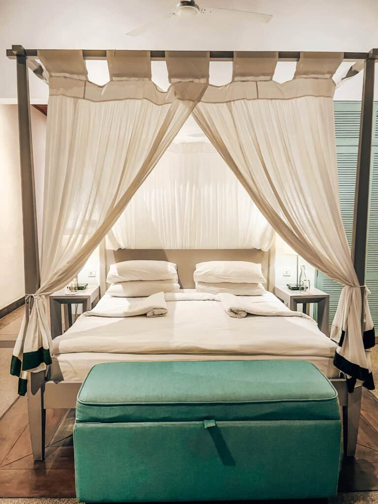 A four-poster bed with white linens and a sheer canopy curtain, complemented by a light turquoise bench at the foot of the bed. The room has soft, calming colors, with bedside tables and ceiling fans adding to the serene atmosphere.