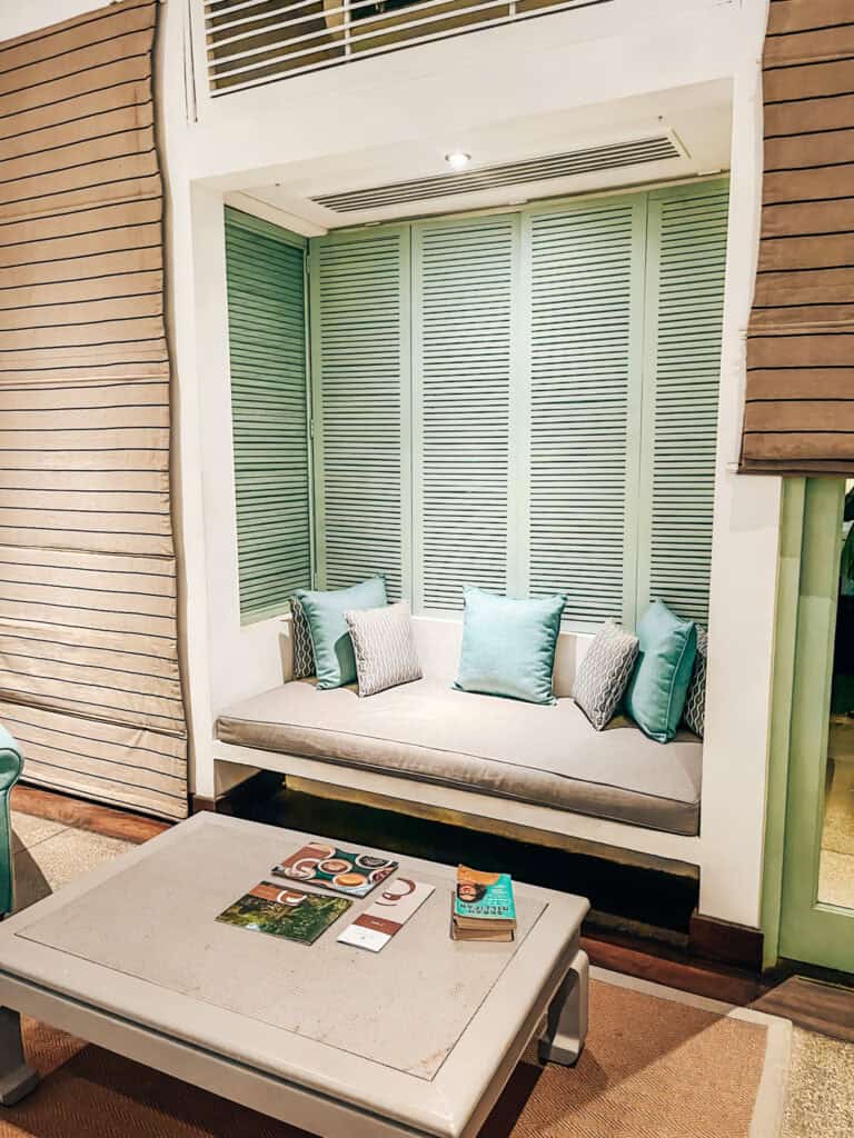  A sitting area featuring a built-in daybed with light green shutters, decorated with throw pillows. A low coffee table in the center holds books and magazines, creating a relaxed and inviting space.