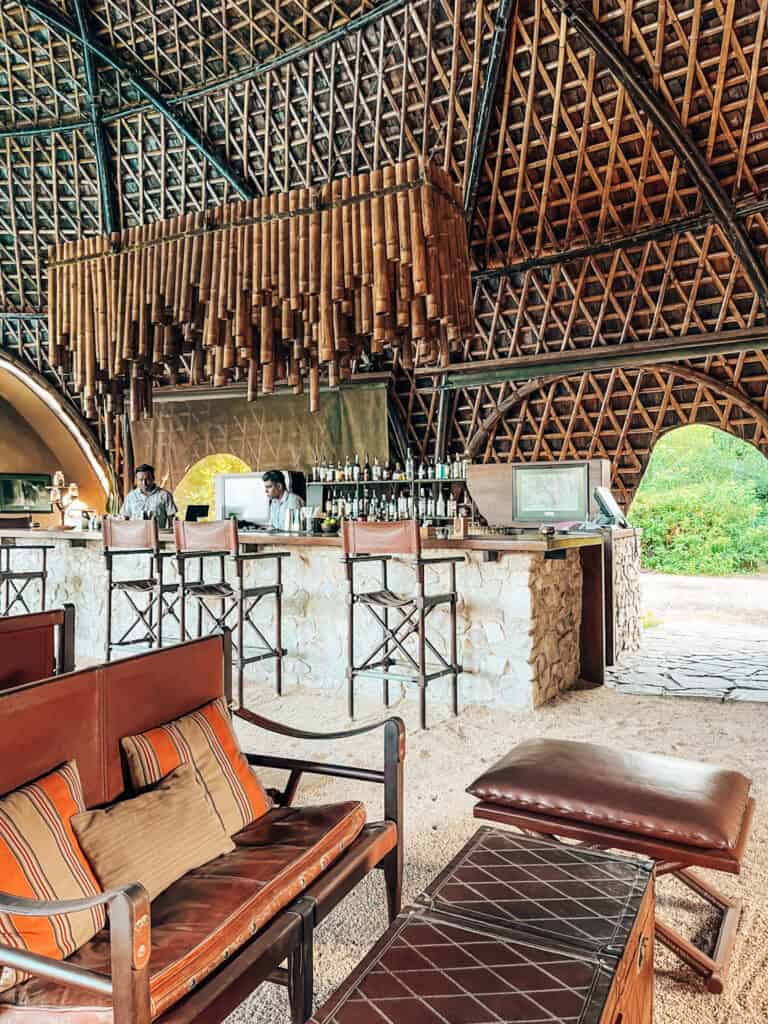 An open-air bar with a woven bamboo ceiling and rustic stone counter. High stools and leather armchairs with striped cushions create a relaxed, luxurious vibe. Bartenders are preparing drinks, with wine bottles and spirits displayed behind the bar.