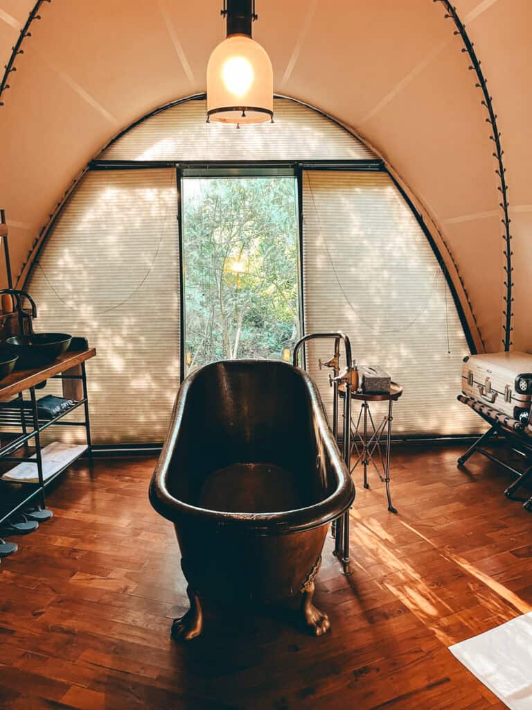 A rustic-style freestanding clawfoot bathtub is centered in a luxury tent with a large arched window overlooking greenery. Warm sunlight filters through, creating a serene atmosphere with natural tones of wood and soft lighting.