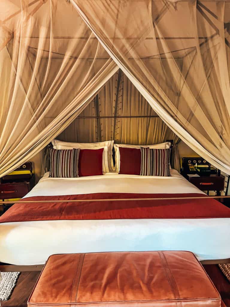 king-sized bed with crisp white linens, red accents, and striped pillows sits under a canopy of sheer fabric, creating a warm, inviting ambiance in a safari-style luxury tent. A leather bench sits at the foot of the bed, and wooden nightstands with vintage decor flank the space.