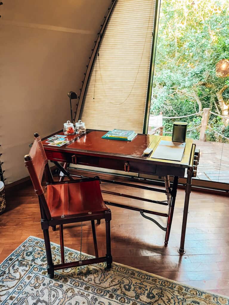 A vintage-style writing desk with a leather chair is positioned near a large window offering a view of lush greenery. The desk holds books, jars, and a small speaker, with a patterned rug adding to the rustic, yet elegant safari tent atmosphere.