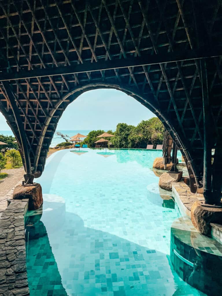 A view through a shaded, arched tunnel structure leading out to a bright, turquoise infinity pool with lounge chairs and umbrellas in the background. The pool extends toward the ocean, framed by greenery and a clear blue sky, offering a tranquil, tropical escape.