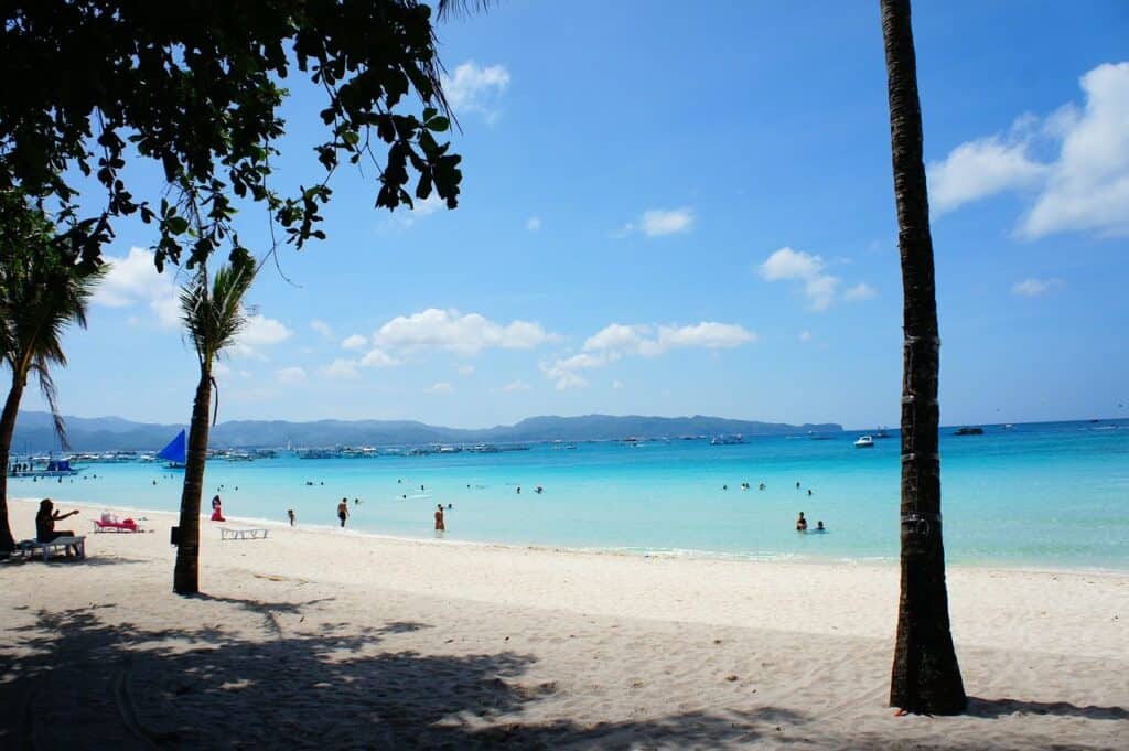 This image shows a beautiful tropical beach with white sand, clear turquoise water, and people swimming and relaxing. Palm trees frame the scene, and in the distance, you can see boats on the water and a mountainous coastline under a sunny sky with scattered clouds. This appears to be a peaceful and scenic beach setting, in Boracay, Philippines.