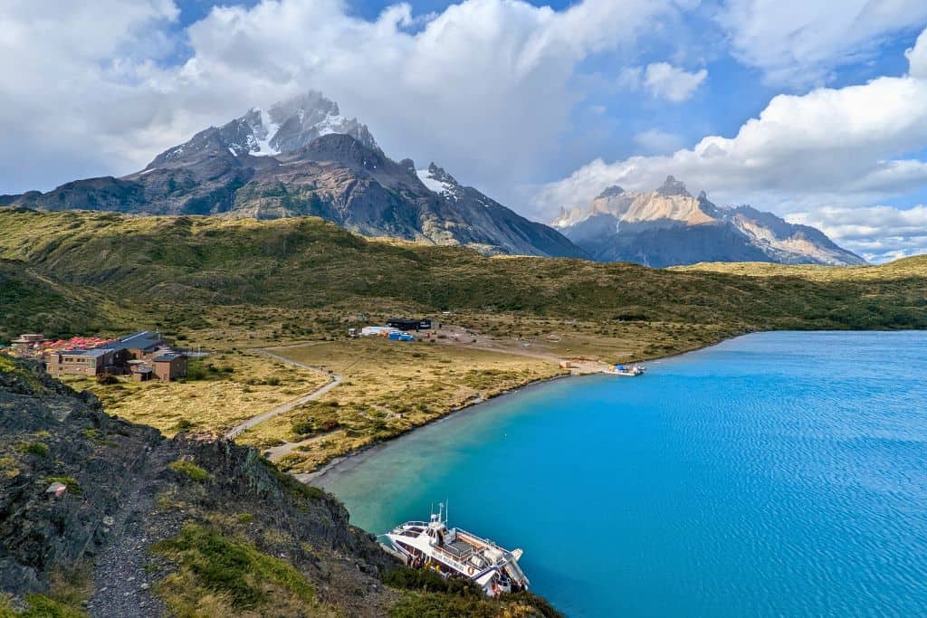 This image captures a stunning view of Torres del Paine in Chilean Patagonia, featuring a vibrant blue lake in the foreground with rugged mountains, partially covered in snow, dominating the background. A small boat is docked by the shore, and several buildings are scattered on the landscape, likely part of a remote lodge or base camp for hikers. The vast, open terrain of grassy plains contrasts with the dramatic peaks and the serene water, highlighting the raw beauty and remote wilderness of this iconic national park.